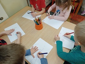 Dzieci siedzą przy stoliku i rysują kredkami wzory na białych kartkach A4.
