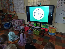 Dzieci siedzą przed monitorem, na którym wyświetlony jest zegar ze wskazówkami, na niebieskim tle. 