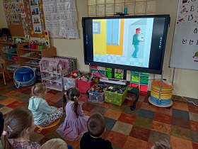 Dzieci siedzą przed monitorem, na którym wyświetlony jest pan, który trzyma w rękach zegar. 