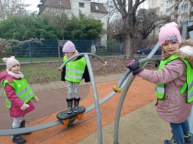 Trzy dziewczynki bawią się na sprzętach na placu zabaw. Mają na sobie kurtki, czapki i rękawiczki, a także zielone, odblaskowe kamizelki.