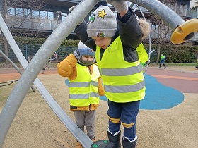 Dwoje dzieci w żółtych kamizelkach odblaskowych bawi się na sprzętach na placu zabaw.