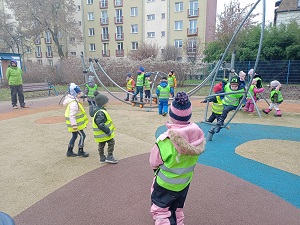 Dzieci w zielonych, odblaskowych kamizelkach bawią się na sprzętach na placu zabaw.