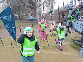 Dzieci w zielonych, odblaskowych kamizelkach bawią się na sprzętach na placu zabaw.