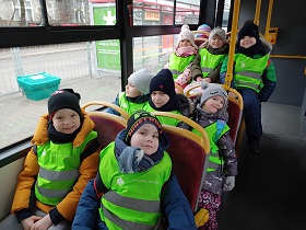 Dzieci w zielonych, odblaskowych kamizelkach siedzą na krzesełkach w autobusie.