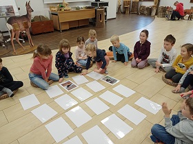 Dzieci siedzą na podłodze w sali. Przed nimi ułożone są białe kartki a4. Dziewczynka odwraca dwie kartki.