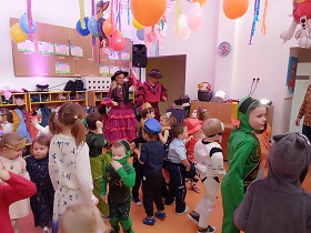 Dzieci w strojach karnawałowych stoją w sali ozdobionej balonami i bibułą. Przed nimi stoi pani i pan w mikrofonami przy buzi. 