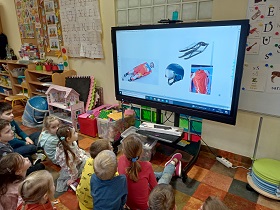 Dzieci siedzą na podłodze przed monitorem. Na monitorze wyświetlany jest slajd przedstawiający saneczkarstwo. Widać na nim kask, sanki, kombinezon oraz zawodnika. 