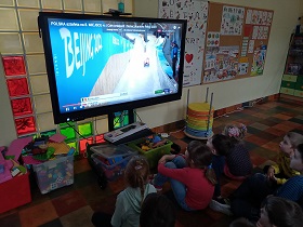 Dzieci siedzą na podłodze przed monitorem. Na monitorze wyświetlany jest film z saneczkarstwem.