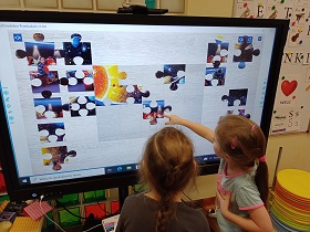 Dwie dziewczynki stoją przy monitorze. Jedna z nich dotyka ekranu, na którym wyświetlone są puzzle.