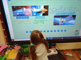 Dziewczynka stoi przy monitorze i rozwiązuje na nim zadanie. Na monitorze widać sondę, rakietę oraz satelitę.