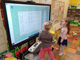 Dwie dziewczynki stoją przy monitorze i rozwiązują zadanie z przeliczaniem elementów.