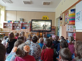 Dzieci siedzą na podłodze przed monitorem, na którym wyświetlany jest film. 