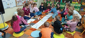 Dzieci wraz z panią w czerwonej bluzce siedzą w kółku na podłodze. Przed nimi leżą białe ilustracje ułożone w jednym rzędzie. Chłopiec w niebieskim swetrze układa coś przy nich. 