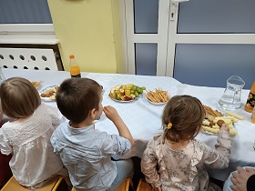 Trójka dzieci: w środku chłopiec, po lewej i po prawej stronie dziewczynki w jasnych sukienkach siedzą przy stole i jedzą poczęstunek.