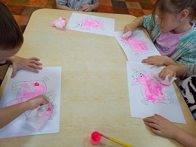 Przy stole siedzi czworo dzieci, na palcach mają owinięte chusteczki zamoczone w farbie, którymi wypełniają kontur świnki.