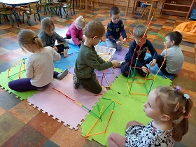 Na matach siedzi ośmioro dzieci, które z kolorowych patyczków układają budowle.