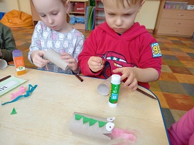 Dwoje dzieci siedzi przy stoliku. Dziewczynka trzyma rolkę po papierze toaletowym i przykleja kolorowy pasek krepiny. Na stoliku leży rolka – smok.