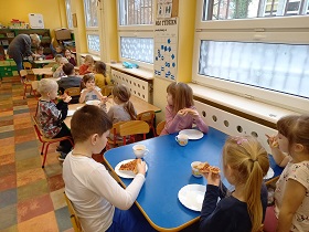 Dzieci siedzą przy stolikach i jedzą pizze, którą mają położoną na talerzykach. 