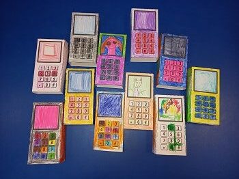Na stoliku leży jedenaście papierowych telefonów komórkowych. Każdy telefon jest inaczej pokolorowany.