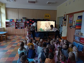 Dzieci siedzą na podłodze przed monitorem, na którym wyświetlana jest mała dziewczynka, która się uśmiecha. Obok monitora stoją dwie panie i dziewczynka. 