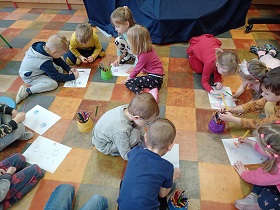 Dzieci pracują w grupach na podłodze. Przed nimi leżą białe kartki papieru i kredki.