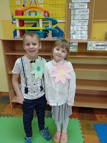 Dziewczynka i chłopiec ubrani na galowo pozują do zdjęcia i się uśmiechają. Na swoich bluzkach mają papierowe, duże kwiaty.