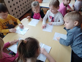 Dzieci siedzą przy stoliku i palcami przesuwają pastę do zębów po papierowych ząbkach wsadzonych w koszulki.