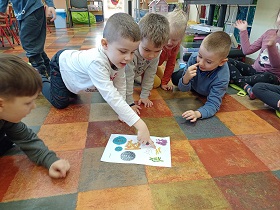 Dzieci siedzą na podłodze i wskazują palcami na kartkę z narysowanymi bakteriami i wirusami. 