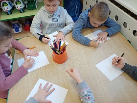 Dzieci siedzą przy stolikach i odrysowują swoje dłonie. Na środku stołu stoi pudełko z kredkami. 