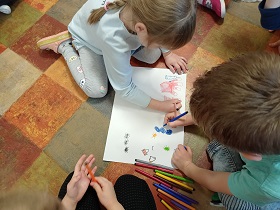 Dziewczynka i chłopiec rysują na kartce papieru mazakami. 