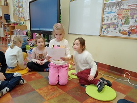 Dziewczynka w różowych spodniach i białej bluzce trzyma w rękach kartkę, na którą patrzy. Obok niej siedzą dziewczynki, które na nią spoglądają. 