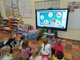 Dzieci siedzą na kolorowych poduszkach przed monitorem, na którym wyświetlane są zdjęcia w białych kółkach na niebieskim tle. 