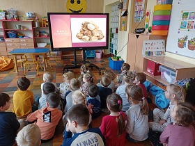 Dzieci siedzą na podłodze przed monitorem, na którym wyświetlane są pączki oraz napis tłusty czwartek. 