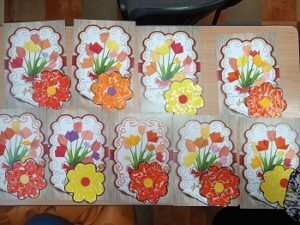 Na stoliku znajdują się laurki w postaci kartek z narysowanymi wazonami i doklejonymi kwiatkami oraz z dużym kwiatem.