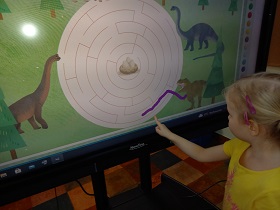 Przed tablicą multimedialną stoi dziewczynka, która rysuje drogę dinozaura do gniazda.