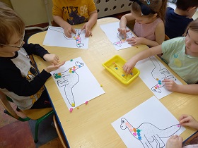 Przy stoliku siedzi piątka dzieci, każde wykleja rysunek dinozaura kulkami plasteliny.