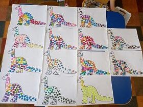 Na stole leży czternaście kartek z rysunkiem dinozaura, które wyklejone są kolorowymi kulkami plasteliny.