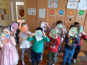 W sali stoją dzieci i trzymają przed swoją twarzą pokolorowaną głowę dinozaura.
