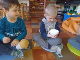 Na podłodze siedzi dwóch chłopców. Chłopiec po prawej stronie trzyma w dłoniach białe jajko.