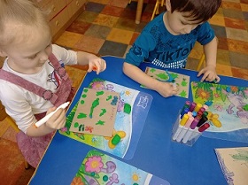 Dwoje dzieci siedzi przy stoliku i rysuje na tekturkach flamastrami.