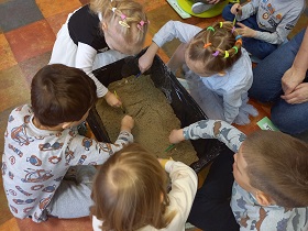 Nad pudełkiem z piaskiem nachylone są dzieci, które pędzelkami przesuwają piasek.