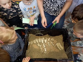 Dzieci siedzą wokół pudełka z piaskiem. Wewnątrz widać szkielet dinozaura.