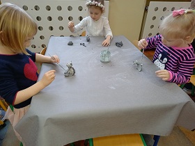 Przy stoliku siedzą trzy dziewczynki. Każda w ręku trzyma pędzelek którym maluje swoją figurkę.