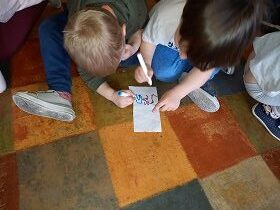 Dwoje dzieci siedzi na podłodze. Przed nimi leży biały prostokąt na którym dzieci rysują flamastrami.