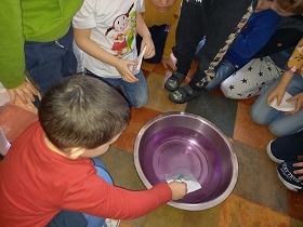 Wokół miski zgromadzone są dzieci. Jedno dziecko wkłada do miski z wodą białą kartkę na której widać rysunek. 