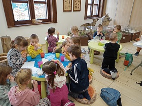 Dzieci siedzą przy zielonych stolikach. Jedzą kanapkę, jabłka i piją herbatę. 
