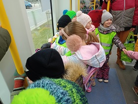 Dzieci w strojach zimowych i zielonych kamizelkach trzymają się poręczy w tramwaju. 
