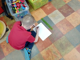 Chłopiec w czerwonej koszulce zapisuje coś na białej kartce, obok której leży linijka. 