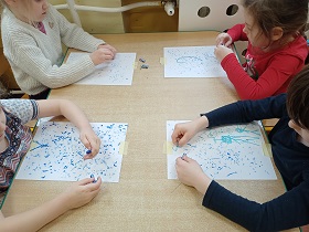 Dzieci siedzą przy stoliku i niebieskimi pastelami rysują na kartkach papieru kropelki deszczu.