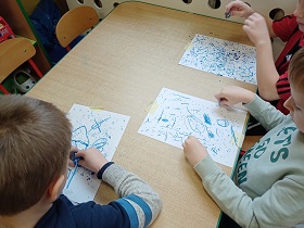 Dzieci siedzą przy stoliku i niebieskimi pastelami rysują na kartkach papieru kropelki deszczu.